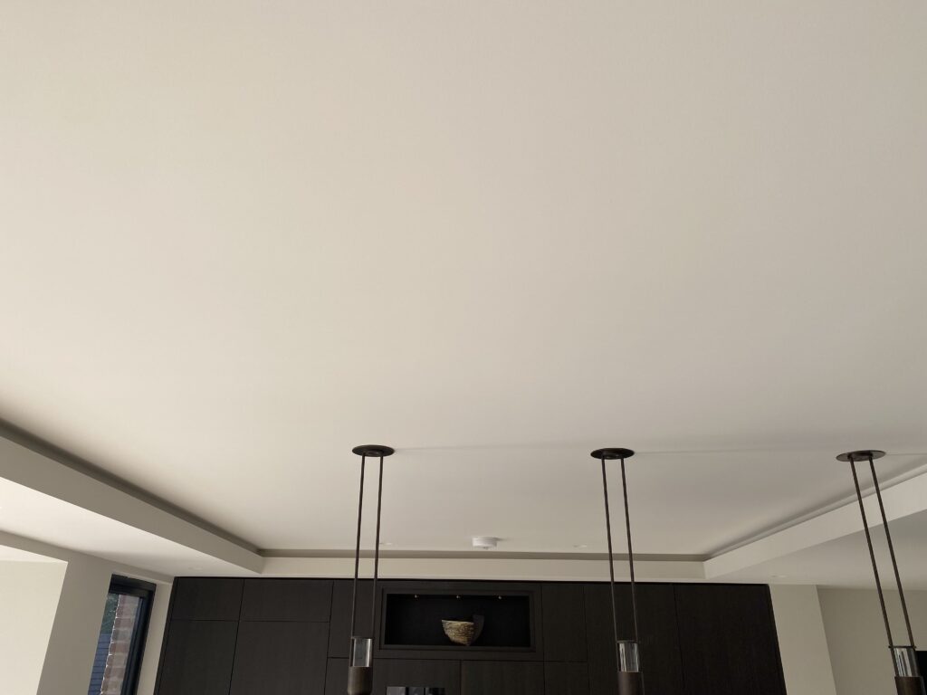 A minimalist ceiling with three modern pendant lights hanging down; dark cabinets and a decorative bowl on a shelf are visible below. The room is bright with natural light coming from a window on the left.