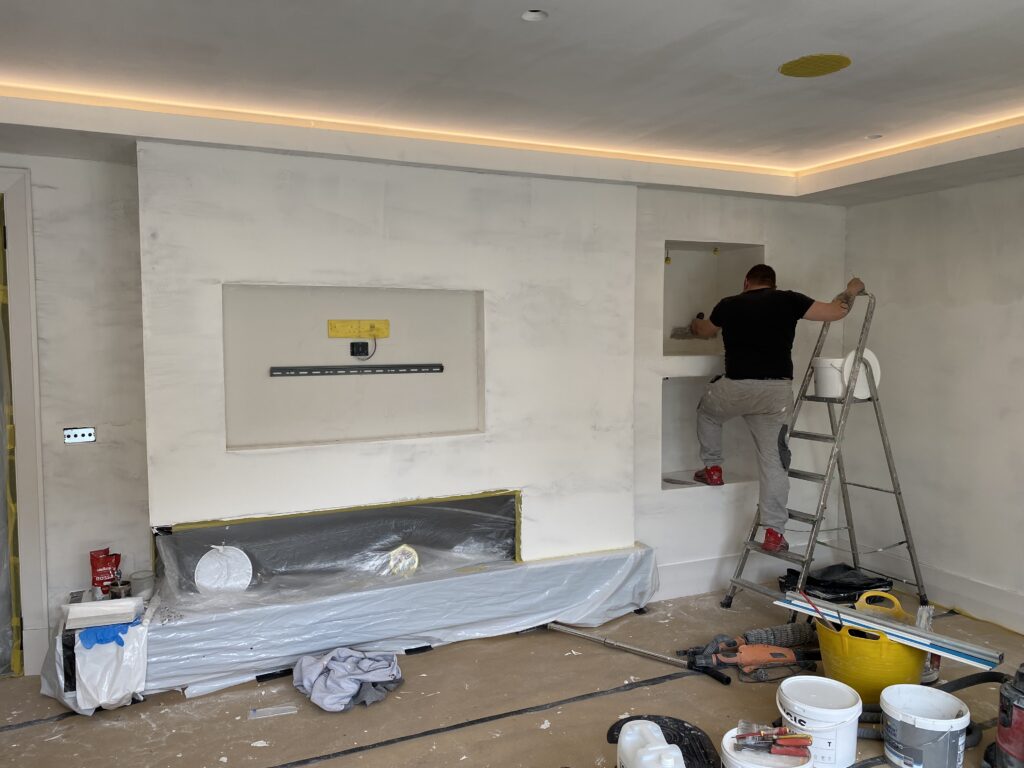 A person standing on a ladder works on built-in shelves in a partially finished room. Tools and supplies are scattered on the floor, and the walls and niche areas are primed and unpainted.