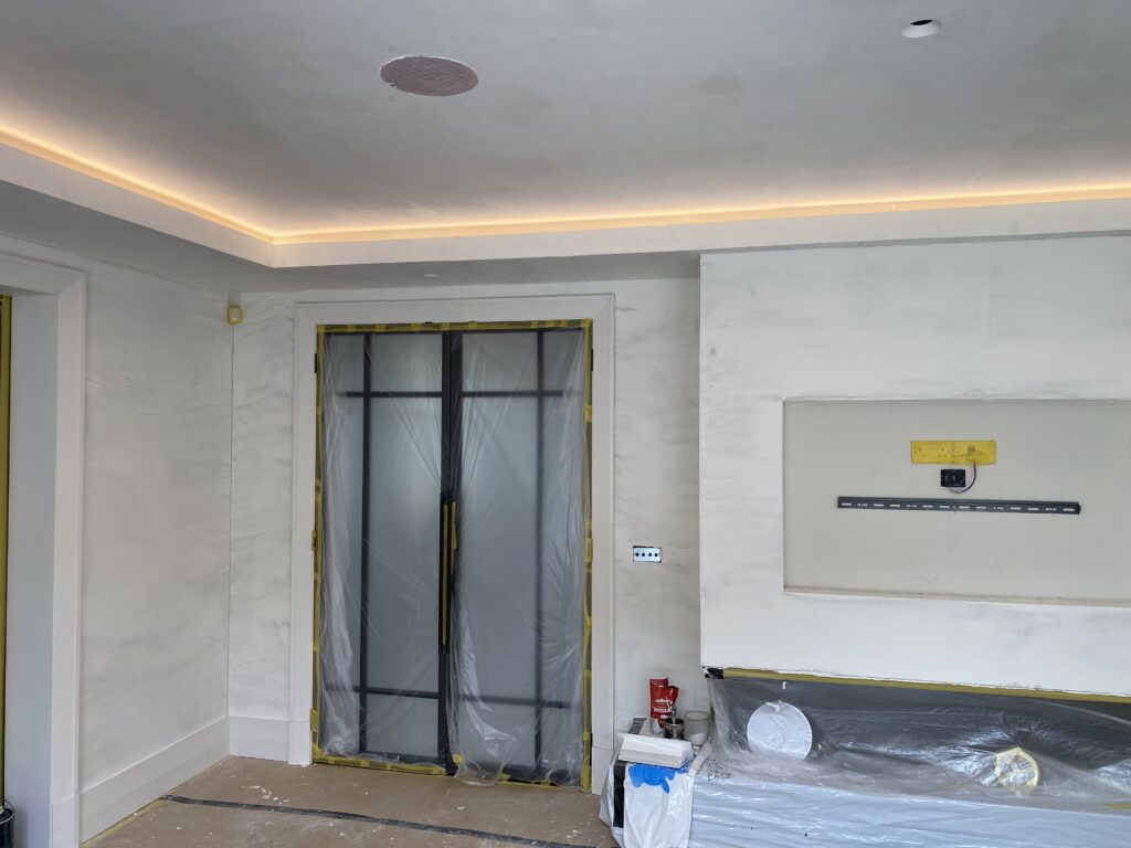 A room under renovation with white walls, plastic covering a door, taped-off areas, recessed ceiling lights, and construction materials on the floor and against the wall.