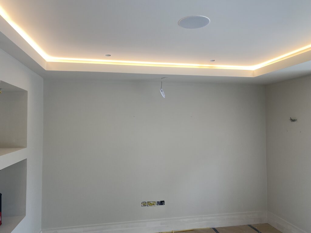 A room with plain white walls, recessed ceiling with hidden LED strip lighting, and some electrical wires and outlets exposed. Built-in wall shelves are on the left, and the floor appears unfinished.