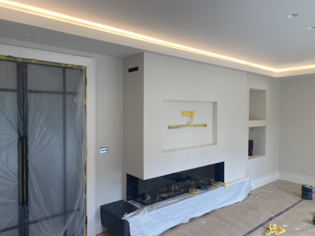 A modern living room under renovation with a built-in fireplace, recessed wall niches, LED ceiling lighting, and protective plastic covering on the floor and doorway. Tools and materials are scattered around.