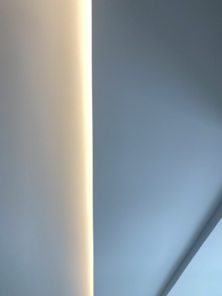 Soft, indirect light creates a glowing line along a smooth, pale wall, dividing the image between a warm, off-white area on the left and a cool blue-gray area on the right. The scene is minimalist and abstract.