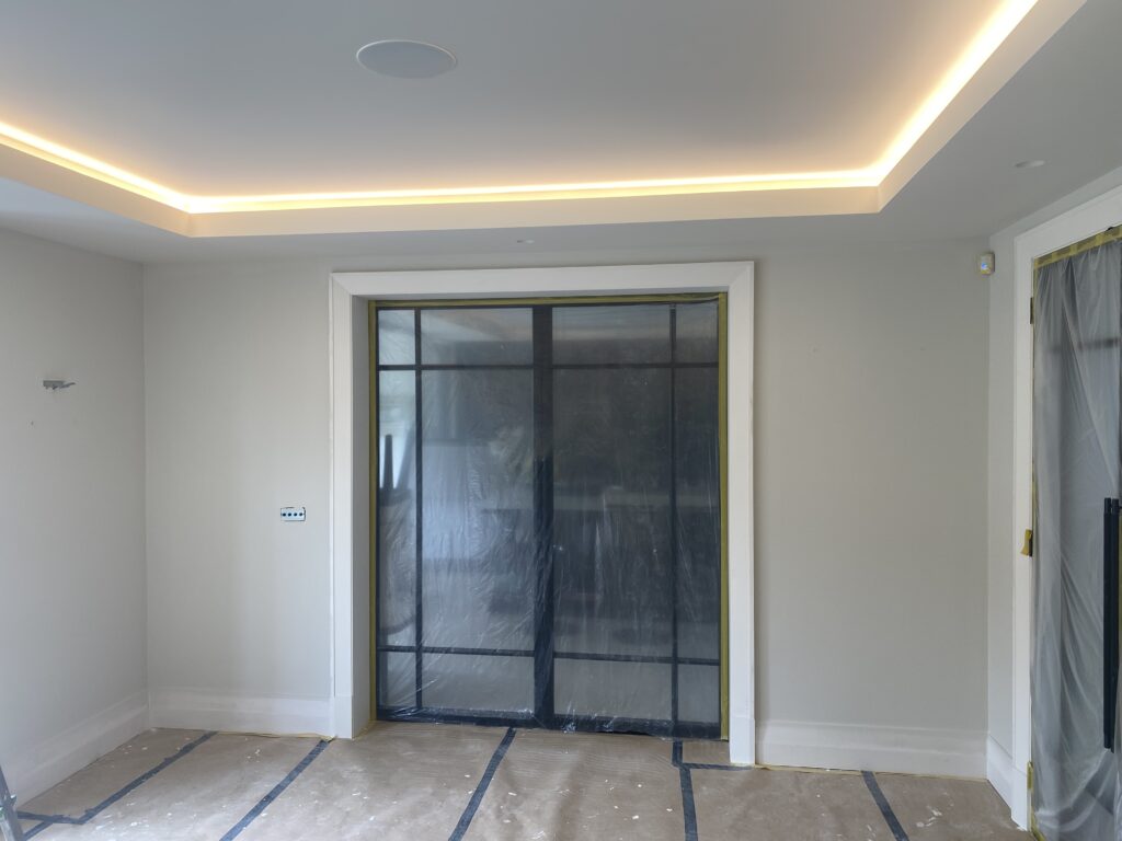 A modern, unfinished room with recessed ceiling lights, a large glass sliding door covered with plastic, and protective coverings on the floor and adjacent door. Walls are light-colored and the room appears to be under renovation.