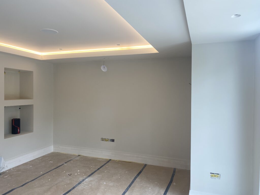 A room under renovation with bare walls, recessed ceiling lights, two open wall niches on the left, exposed wiring, and protective covering on the floor. The space is lit by natural light from an unseen window.