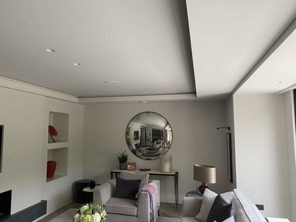 A modern living room with neutral walls, a large round mirror, a grey sofa, a beige armchair, a table lamp, decorative shelves with red accents, and a glass door letting in natural light.