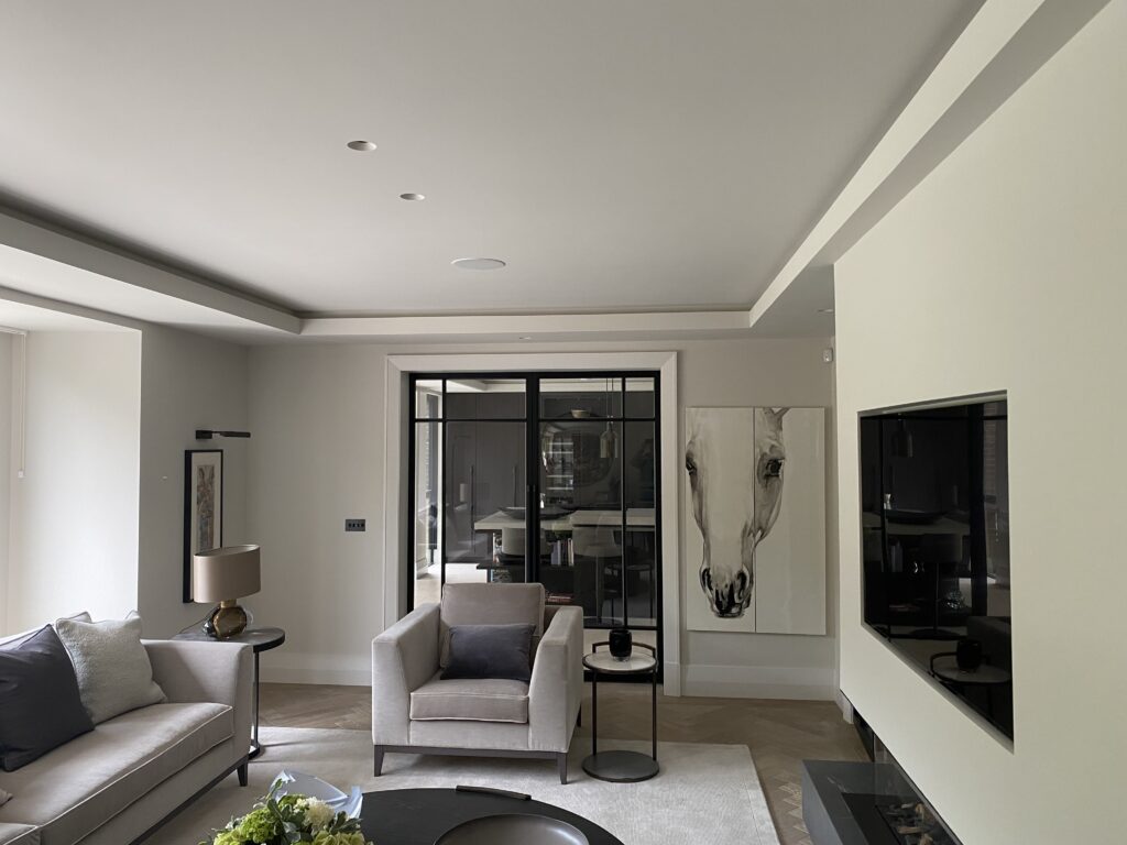 Modern living room with neutral tones, featuring a sofa, armchair, side tables, large wall art of a horse, and a flat-screen TV. Glass doors at the back reveal another room, adding depth to the space.