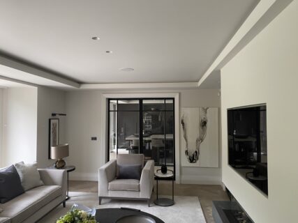 Modern living room with neutral tones, featuring a sofa, armchair, side tables, large wall art of a horse, and a flat-screen TV. Glass doors at the back reveal another room, adding depth to the space.
