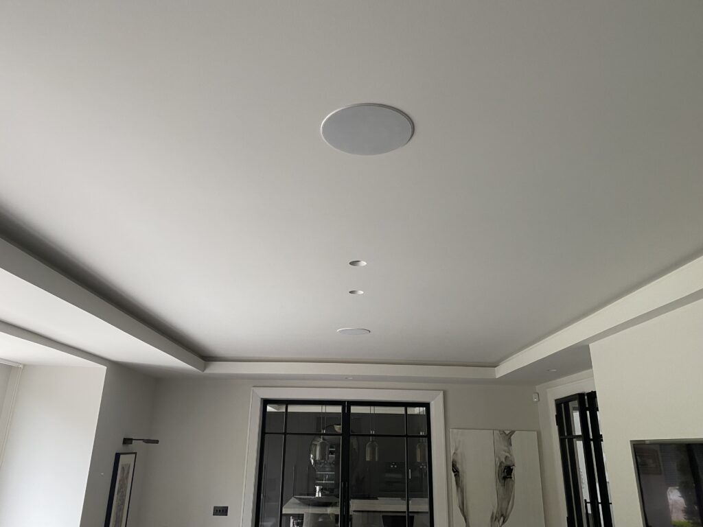 A modern room ceiling with two circular built-in speakers and three recessed spotlights. The room has white walls, large windows, and framed artwork visible on the walls.