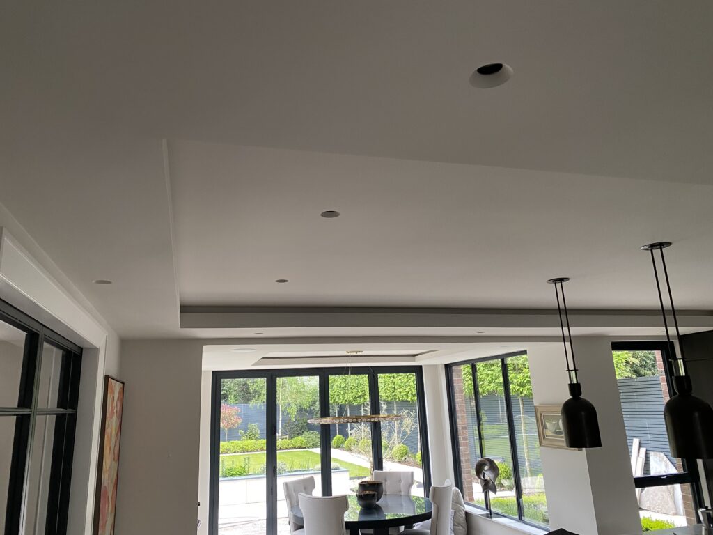 Bright dining area with large windows overlooking a green garden. There is a round table with white chairs, modern pendant lights, and artwork on the wall, creating a contemporary and airy atmosphere.