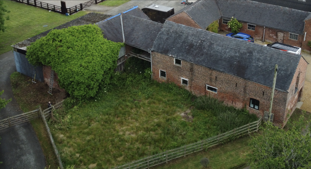 An aerial view of a brick farm building with a partially overgrown courtyard, surrounded by a wooden fence. One side of the building is covered in dense green ivy. There are cars and other buildings nearby.