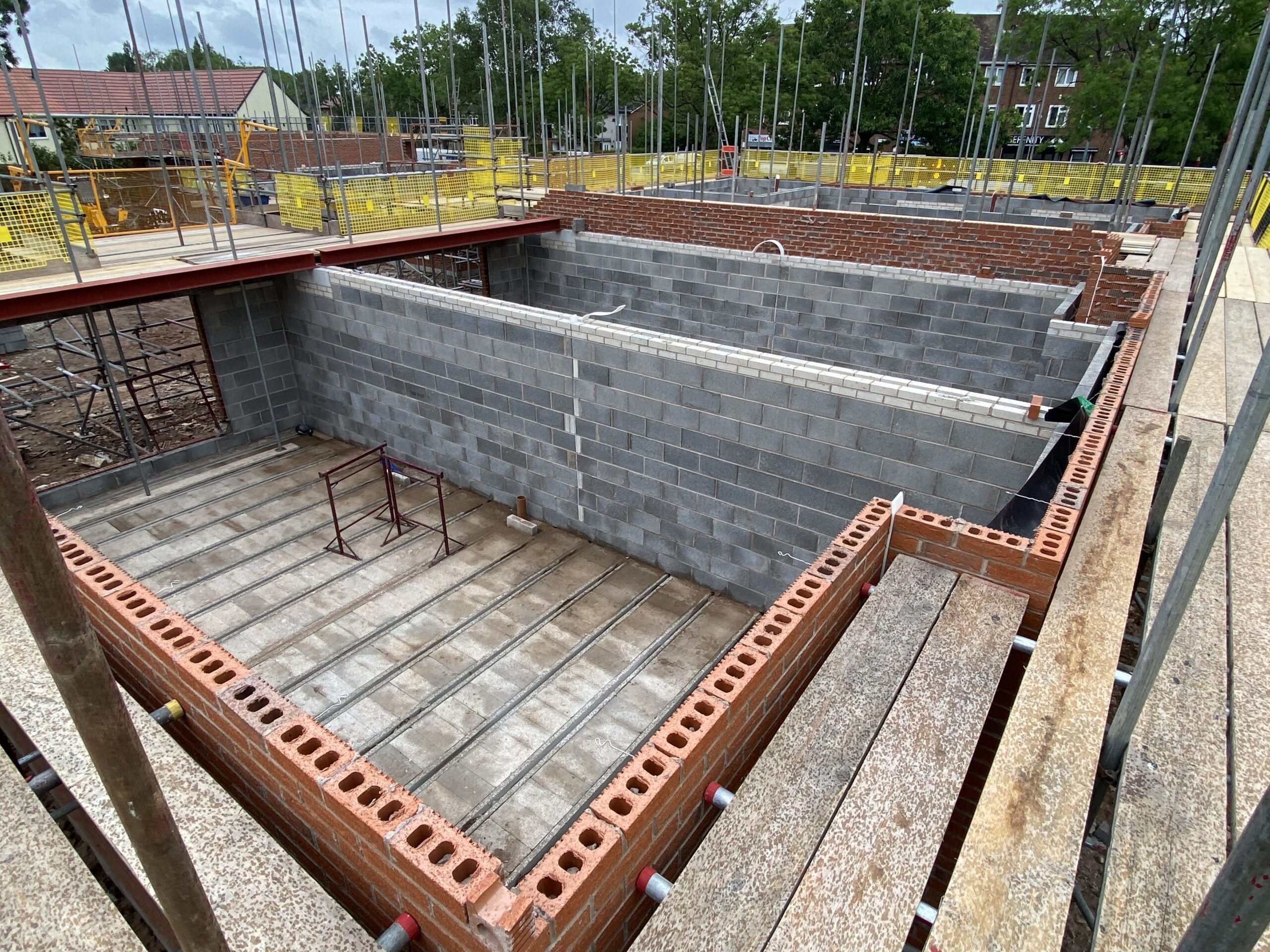 View of a construction site showing the foundation and lower brick walls of a building. Concrete blocks form interior walls, and red bricks form outer walls. Metal rods and scaffolding surround the site.